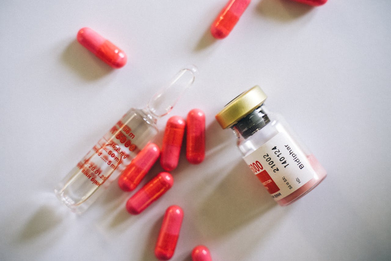 Flat lay of medication bottle, glass vial, and red capsules on a white surface.